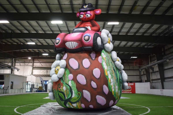 Buggy Bear, a giant inflatable character with a red cap, pink spotted body and green flowers, standing on turf inside a large warehouse