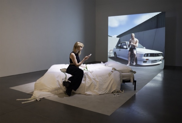 The artist sits on a bed draped in white sheets in a dark room, looking at her phone, in front of a large screen showing a man leaning on a white car