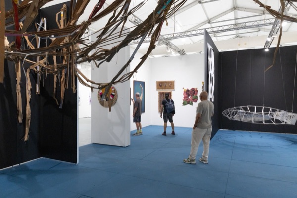 Visitors walk across a blue floor between gallery walls, under a hanging sculpture of torn cardboard strips, with a suspended white wire form to the right