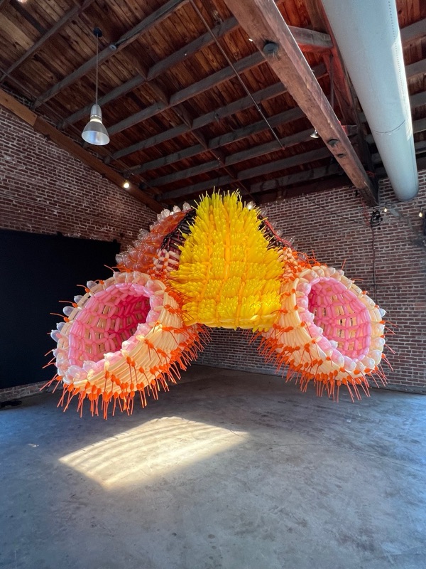 A large hanging sculpture of ribbed coral, orange and pink forms in a gallery with brick walls