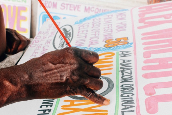 Nurse’s hand, holding a fine brush, painting red letters on a sign crowded with pink, blue and black lettering