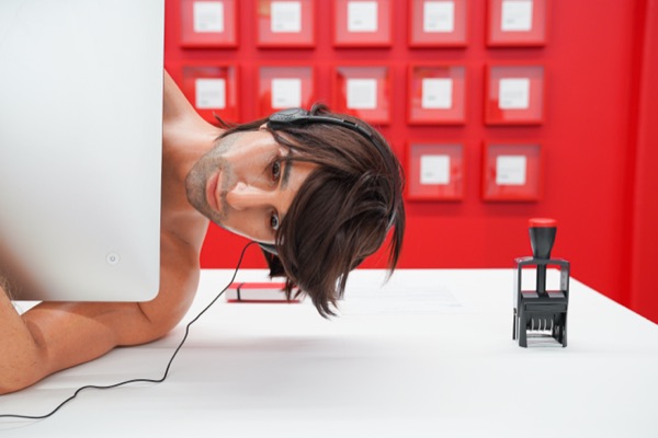 A lifelike sculpture of a man in headphones, his head resting on a white desk, in front of a red wall of framed certificates