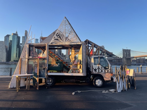 The House Our Families Built, a house-shaped sculpture with cutaway walls on the back of a truck, on a waterfront at dusk with the Manhattan skyline behind