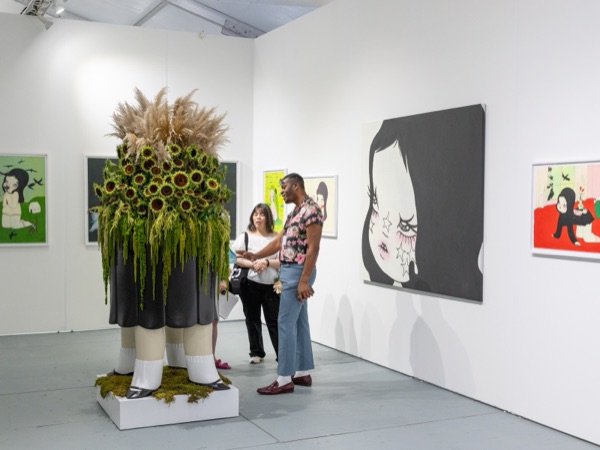 Visitors in a gallery booth beside a tall sculpture crowned with sunflowers
