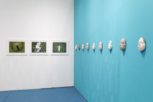 Three framed photographs of a figure in white lying on grass, beside a turquoise wall hung with a row of cast faces