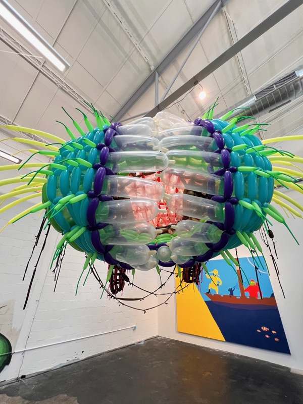 A green, blue and pink sculpture suspended in the corner of a gallery