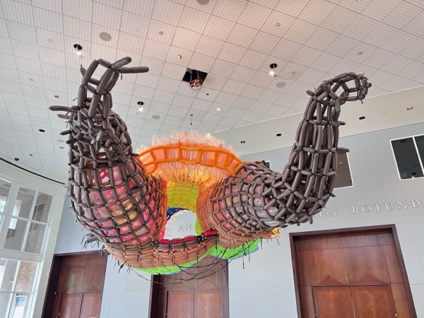 Two dark ribbed forms with orange edges hanging from the ceiling of a white gallery