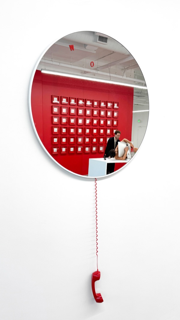 A round mirror on a white wall, with a red telephone receiver hanging below it