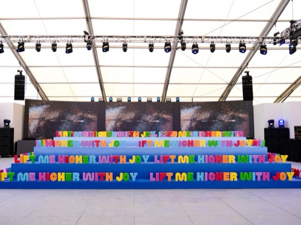 A tiered rainbow installation spelling out Lift Me Higher With Joy, inside a white tent