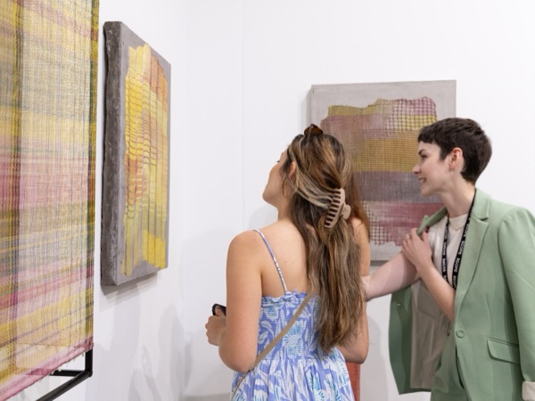 Two visitors talking in front of woven and painted works in a gallery booth