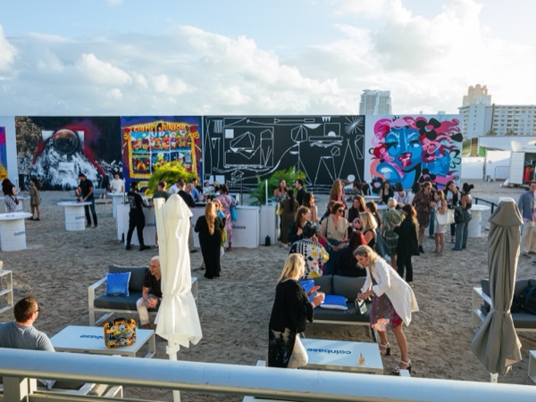 A beach lounge on the sand, with seating, sculptures and a long painted mural wall under a blue sky