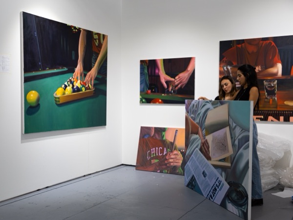 A gallery booth with paintings of a pool table and hands, some still leaning against the wall