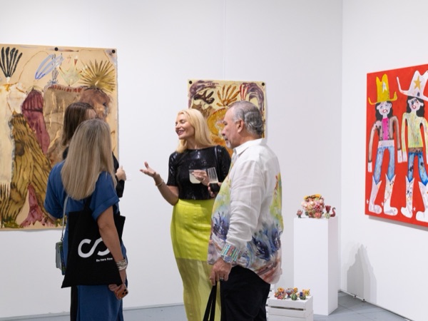 Three visitors in conversation in a gallery booth hung with paintings