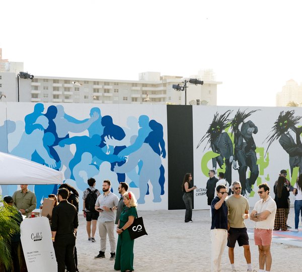 Visitors gather on the sand in front of a long wall of large painted murals, with a food stand at left and apartment buildings behind