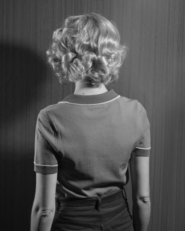 Hella Hammid’s Back by Diana Sosnowska: a black-and-white photograph of a woman seen from behind, with curled blonde hair and a short-sleeved knit shirt, against a wood-grain wall