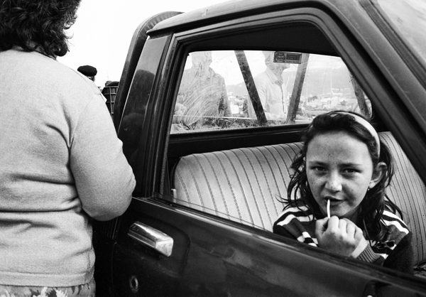 A girl holding a lollipop looks out of a car window while a woman stands beside the open door