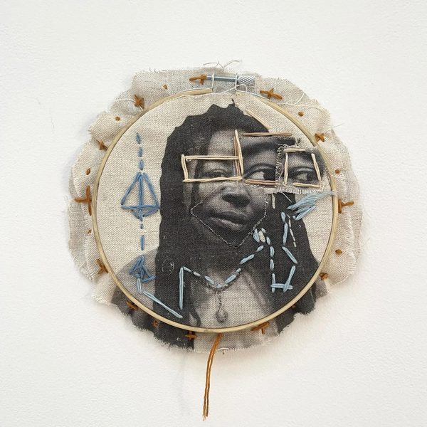 A fabric work in an embroidery hoop: a black-and-white portrait of a woman printed on cloth, stitched over with blue and gold thread, with frayed edges